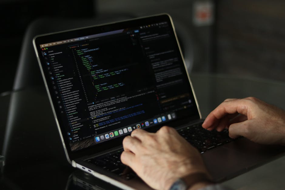 A close-up view of a person’s hands working on a laptop placed on a dark surface, with the screen displaying lines of colorful code against a black background. The person appears to be engaged in co