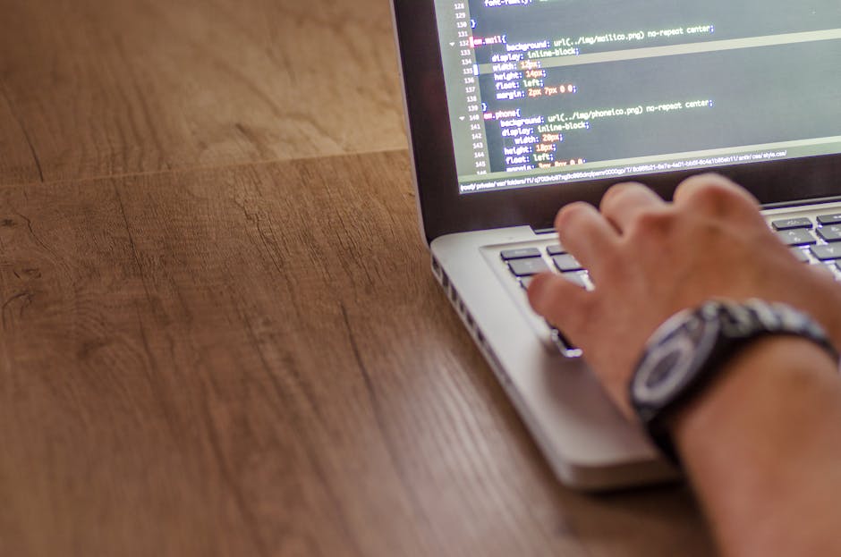 A close-up view of a person’s hands working on a laptop placed on a dark surface, with the screen displaying lines of colorful code against a black background. The person appears to be engaged in co